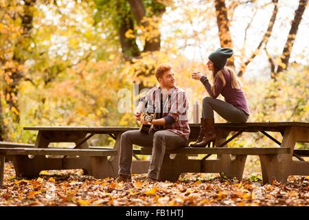 Jeune couple qui joue de la guitare sur des bancs de pique-nique en forêt d'automne Banque D'Images