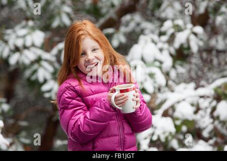 Fille aux cheveux rouge en face d'arbres couverts de neige, portant manteau matelassé rose holding mug à la route smiling Banque D'Images