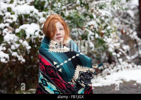 Fille aux cheveux rouge en face d'arbres couverts de neige, enveloppé dans une couverture motif aztèque looking at camera Banque D'Images