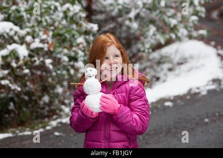 Fille aux cheveux rouge en face d'arbres couverts de neige, le port de gants et veste matelassée rose holding snowman à la route smiling Banque D'Images