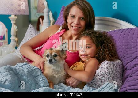 La mère et la fille et le chien de câlins au lit à la caméra en souriant Banque D'Images