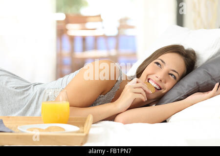 Happy girl eating cookies diététiques au petit-déjeuner et de penser à la plus étendue sur le lit à la maison Banque D'Images