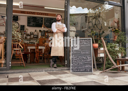 Tirer sur toute la longueur d'un barista jeunes debout dans porte d'un café. Jeune homme avec barbe portant un tablier, debout avec son Banque D'Images