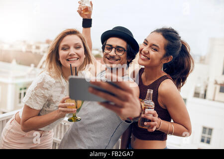 Trois jeunes amis taking self portrait with smart phone pendant une partie. Heureux jeune homme et femme en tenant à selfies par sur le toit Banque D'Images