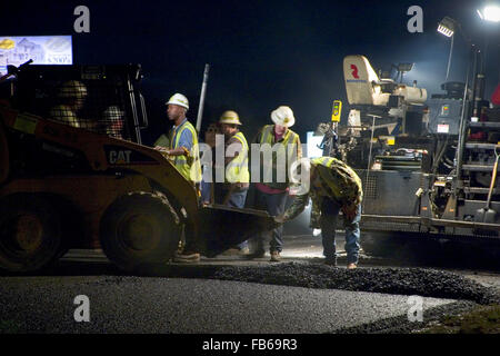 Les travailleurs se tiennent derrière un épandeur d'asphalte mélangé à chaud mise en route inter-États de travailler de nuit. Banque D'Images