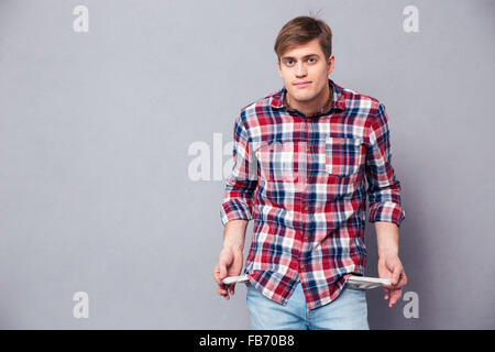 Pauvre beau jeune homme en chemise à carreaux et jeans montrant poches vides sur fond gris Banque D'Images