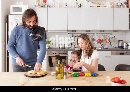 Père hacher les légumes avec la mère et bébé fille jouant dans la cuisine Banque D'Images