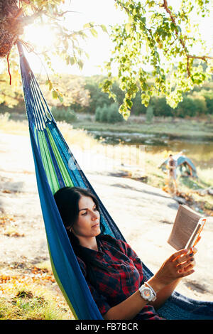 Young woman reading book while lying on hammock in forest Banque D'Images