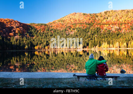 Jeune couple assis sur un banc, sur la rive d'un lac.l'eau est en raison de la forêt d'automne Banque D'Images