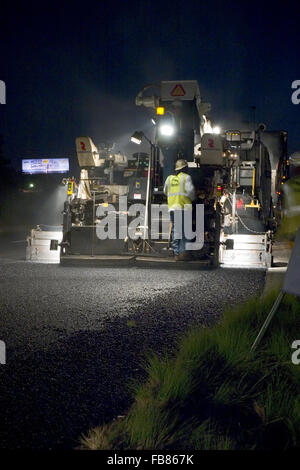 Les travailleurs se tiennent derrière un épandeur d'asphalte mélangé à chaud mise en route inter-États de travailler de nuit. Banque D'Images