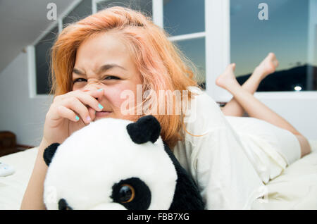 Jolie jeune femme confortablement allongé sur un lit blanc et hugging stuffed animal panda avec de grandes fenêtres en arrière-plan Banque D'Images