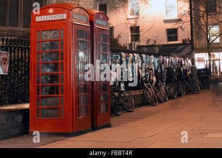 Téléphone ancien style rouge boîtes de téléphone à Cambridge avec affiches sur clôture de fer et des vélos à l'avant, en début de soirée, Faible éclairage Banque D'Images