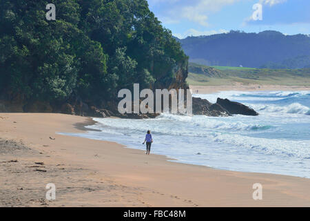 Femme marchant le long de plage de Hot Water -Nouvelle-Zélande Coromandel , avec les vagues se brisant sur la plage à côté d'elle et des buissons . Banque D'Images