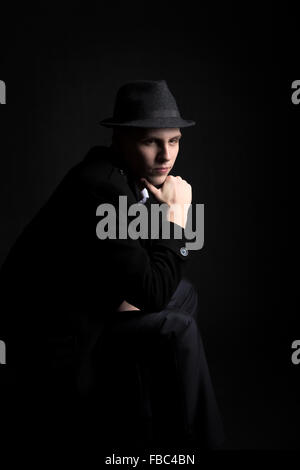 Touche Bas shot de mystérieux jeune homme portant un chapeau et un manteau assis dans l'obscurité avec regard pensif Banque D'Images