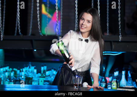 Belle cheerful brunette barman fille en chemise blanche et noeud papillon noir, service de l'alcool boire au comptoir du bar, holding bottl Banque D'Images