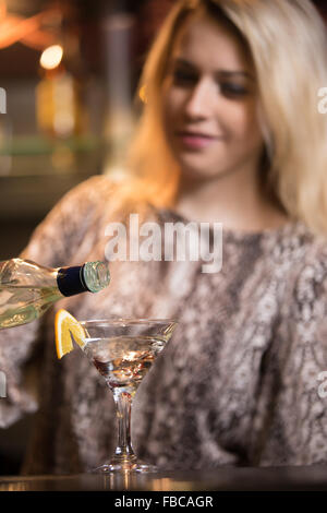 Smiling blonde female bartender préparer l'alcool verre au bar comptoir, tenant en main la bouteille, verser le verre au verre à cocktail, Banque D'Images