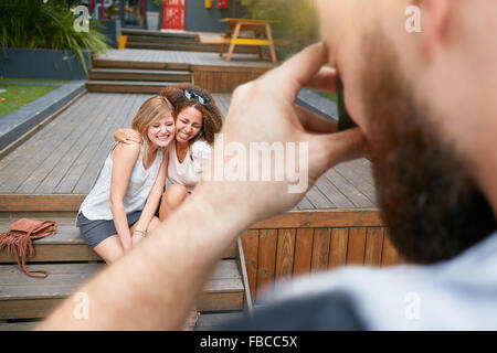 Deux jeunes femme siégeant ensemble souriant et posant pour une photographie. L'homme à prendre des photos de ses amis. Banque D'Images