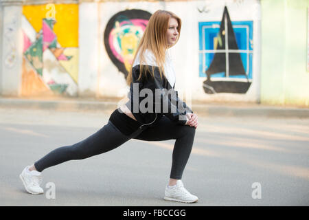 Runner girl with earphones in sportswear faisant des exercices sur une jambe avant de footing matinal sur la rue en face de mur avec Banque D'Images