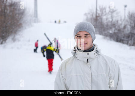 Beau jeune homme sur une piste de ski standing smiling at the camera avec les skieurs et snowboarders en arrière-plan Banque D'Images