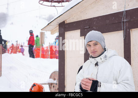 Beau jeune homme jouissant d'un café d'hiver comme il est à côté d'une cabane à une station de ski d'hiver avec les touristes en arrière-plan. Banque D'Images