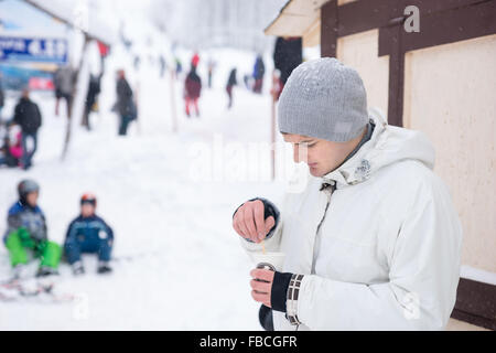 Beau jeune homme de boire du café en hiver qu'il se tient à l'extérieur dans une station de ski en remuant le sucre, haut du corps vue rapprochée avec d'autres personnes comme un flou dans la distance. Banque D'Images