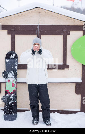 Jeune homme avec son snowboard à boire une tasse de café à emporter pour réchauffer comme il se tient dans la neige en face d'une hutte dans un lieu de villégiature. Banque D'Images