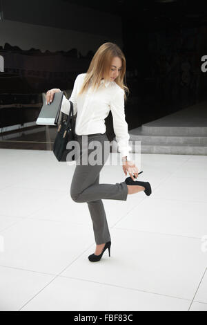 Belle jeune femme en chemise blanche style office holding document noir Sac en cuir, dossier et toucher sa chaussure à haut talon, fe Banque D'Images