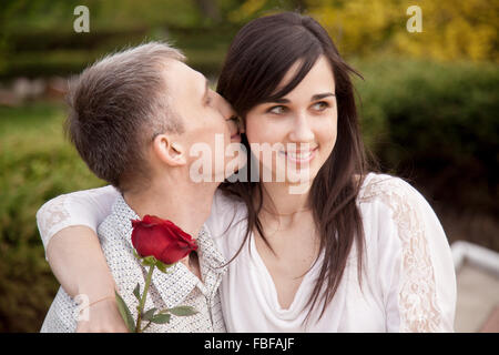 Portrait de couple avec rose rouge sur date dans parc, jeune homme embrassant sa petite amie dans son oreille alors qu'elle est assise sur son tours w Banque D'Images