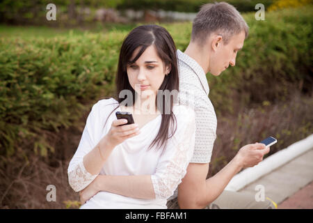 Young Caucasian couple sur date, assis dos à dos sur le banc de parc holding cellphones en mains, à l'aide de l'app, faisant appel, messagin Banque D'Images