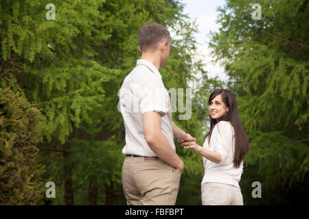 Jeune femme brune en maillot blanc et un jean menant son amoureux à la main dans le parc, regardant avec joyeux sourire adorable, fol Banque D'Images