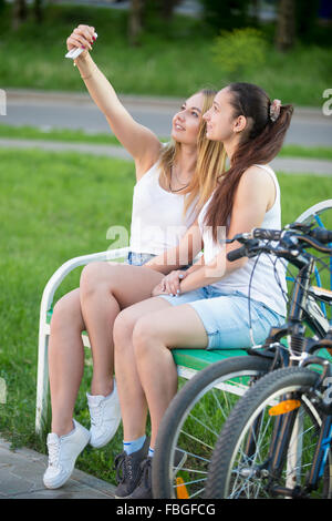 Voir le profil de two cute smiling belle cycliste adolescentes portant des vêtements décontractés assis sur banc de parc sur journée d'été, p Banque D'Images