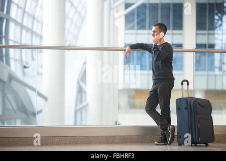 Jeune homme séduisant en 20s debout à côté de mur en verre moderne aéroport terminal, talking on smartphone, voyager avec des bagages Banque D'Images