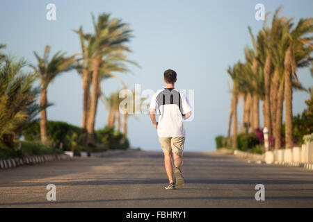 Jeune homme sportif courir loin de l'appareil photo, faire du jogging sur le sunny summer street dans les tropiques, des palmiers sur le backg Banque D'Images