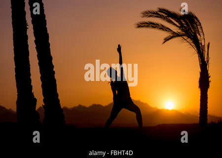 Silhouette de jeune femme pratiquant fitness, yoga ou Pilates au coucher du soleil dans un bel emplacement avec des montagnes et des palmiers, doi Banque D'Images