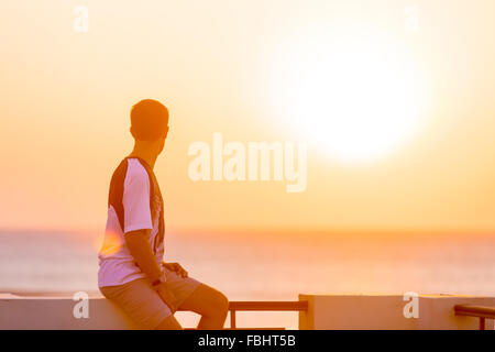 Beau jeune homme assis sur le balcon en fer posent sans souci, profiter du lever ou du coucher du soleil coloré magnifique paysage de mer et f Banque D'Images