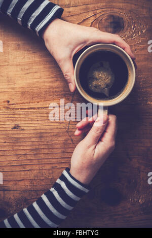 Femme seule à boire du café le matin, top view of female hands holding tasse de boisson chaude sur un bureau en bois, aux couleurs rétro. Banque D'Images
