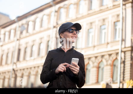 Traveler smiling young woman wearing vêtements décontractés, bonnet et lunettes holding cellulaire, à l'aide de l'app, gps, recherche de directi Banque D'Images