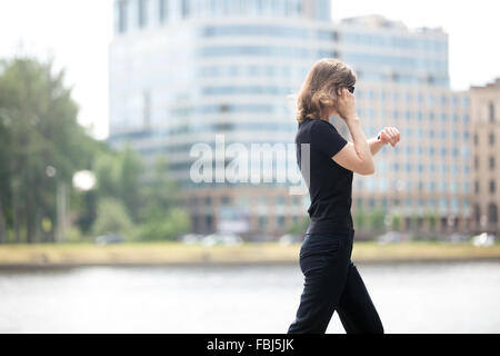 Young business woman walking in hurry, looking at watch, talking on mobile phone sur la rue en face de la ville de blue g Banque D'Images