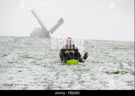 Un homme assis sur un traîneau glisse vers le bas un snow-covered​ colline en face d'un des moulins à vent appelé Jill dans Clayton Clayton, East Sussex, UK. Banque D'Images