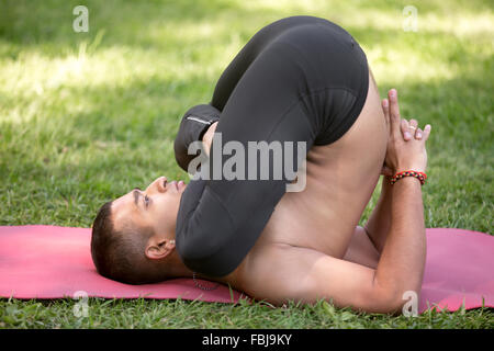 Beau jeune homme indiens travaillant sur mat sur pelouse au parc, faire pindasana dans sarvangasana, variation de la posture de l'embryon, plein Banque D'Images