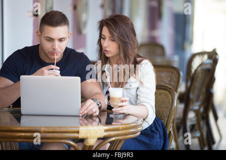 Portrait of young man and woman in casual clothes sitting dans street cafe avec mobilier en rotin de boire du café, à l'aide d'un ordinateur portable, lo Banque D'Images