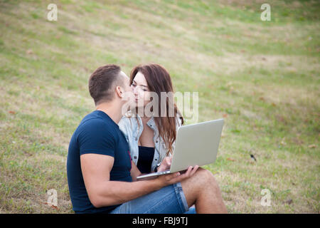 Beau jeune couple sur une date s'embrasser dans le parc, assis sur le gazon sur journée d'été, holding laptop, copyspace Banque D'Images