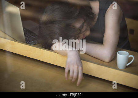 Jeune femme couchée sur le bras sur la table à café en face de l'ordinateur portable avec tasse de café, endormi, fatigué, surmené ou paresseux Banque D'Images