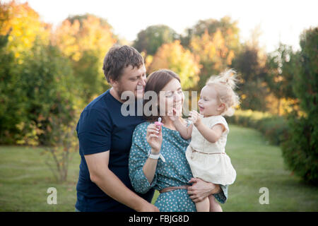 Portrait of happy belle famille de trois personnes à pied dans le parc en automne. Maman et papa carrying baby fille en robe blanche, Banque D'Images