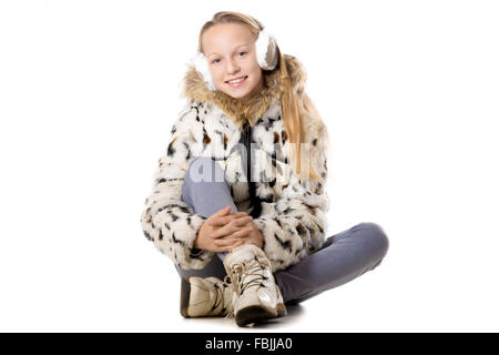 Portrait of happy funny belle adolescente occasionnels portant manteau d'hiver et des cache-oreilles, assis en tailleur, looking at camera Banque D'Images