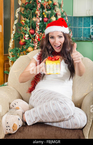 Sweet Young Woman with Christmas Gift Showing Thumbs Up Sitting on Sofa With Santa Hat Smiling Banque D'Images