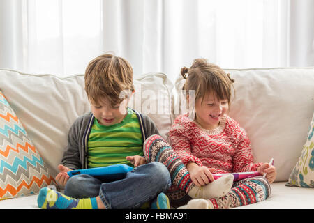 Boy and girl sitting on couch jouant avec les tablettes numériques Banque D'Images