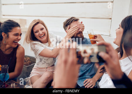 Groupe d'amis s'amuser à une soirée avec l'homme de prendre une photo sur un téléphone intelligent. Les jeunes bénéficiant d'une partie ensemble. Banque D'Images