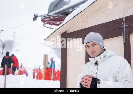 Jeune homme sérieux en manteau et hat holding boisson chaude qu'il s'attendait à un centre de ski Banque D'Images