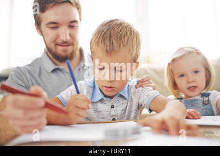 Cute boy dessin avec des crayons de couleur sur fond de son père et sa soeur Banque D'Images